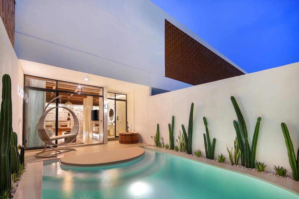A private pool is surrounded by modern landscaping, featuring tall cacti and neatly arranged greenery. A sleek, contemporary structure with large glass doors provides a glimpse into the inviting living area, while soft lighting reflects off the water as twilight casts a serene atmosphere.