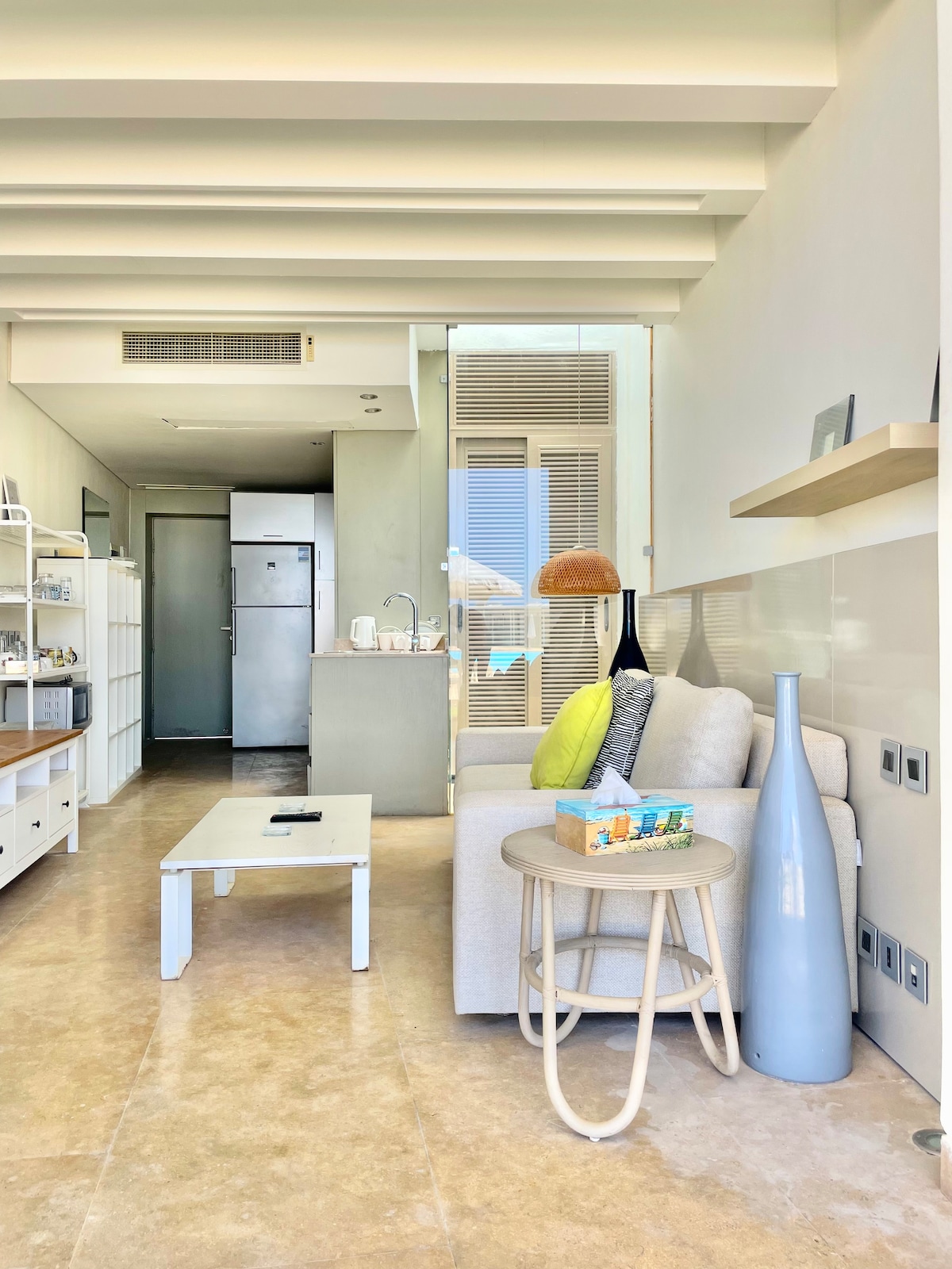A bright living area displays a light-colored sofa paired with a round, wooden side table. A tall, blue vase and a small decorated tray rest on the table. The open kitchen area is visible in the background, featuring modern appliances, including a refrigerator and a sink.