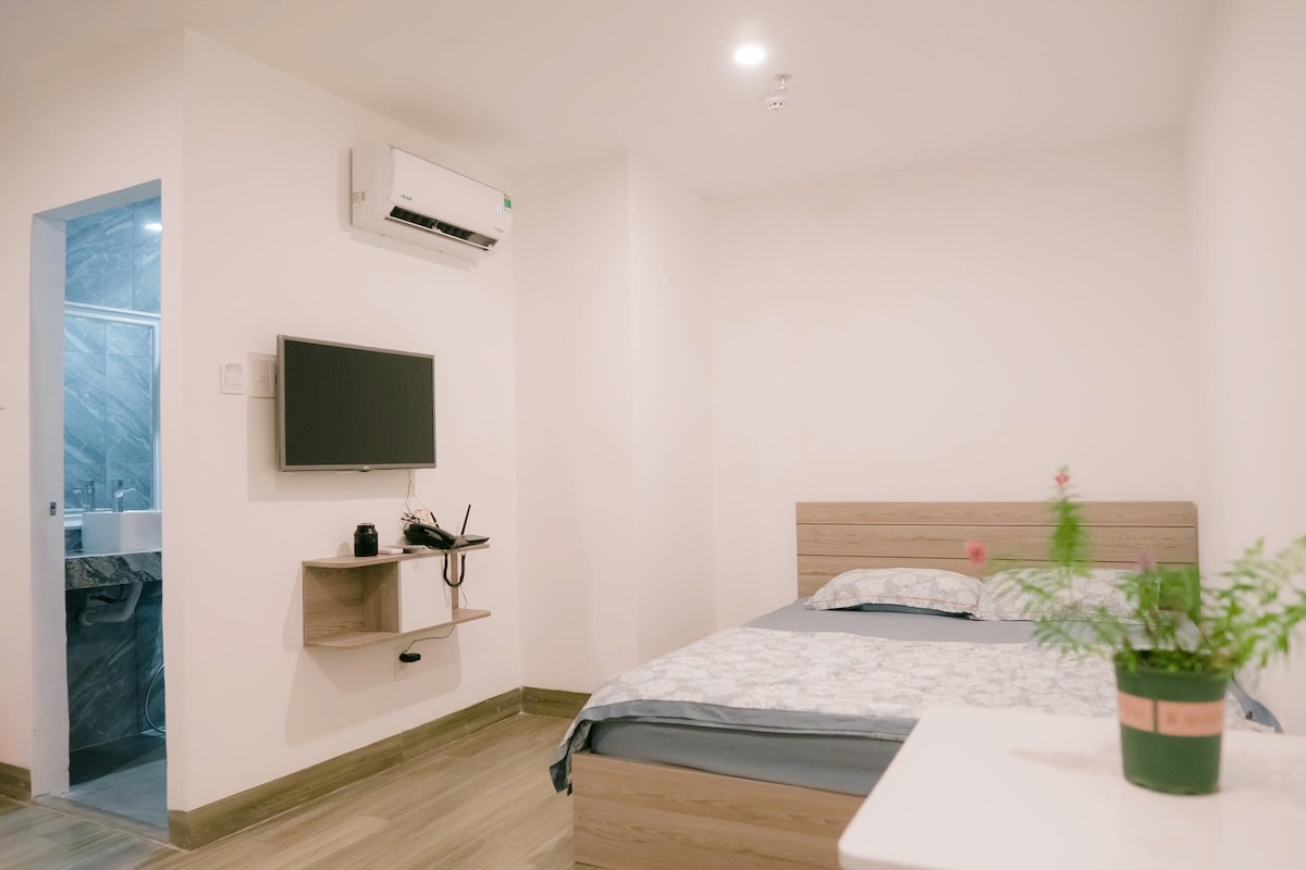 A comfortable sleeping area features a spacious bed with a light-colored quilt. A flat-screen television is mounted on the wall opposite the bed. A small shelf holds a camera, while a potted plant adds a touch of greenery. A bathroom door is visible in the background.