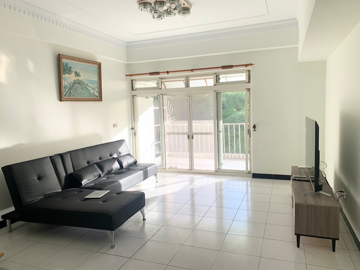A bright living area features a black sofa and a wooden TV stand against a wall. Large glass doors open to a balcony, allowing natural light to fill the space. A framed piece of artwork adorns the wall, adding a touch of interest.