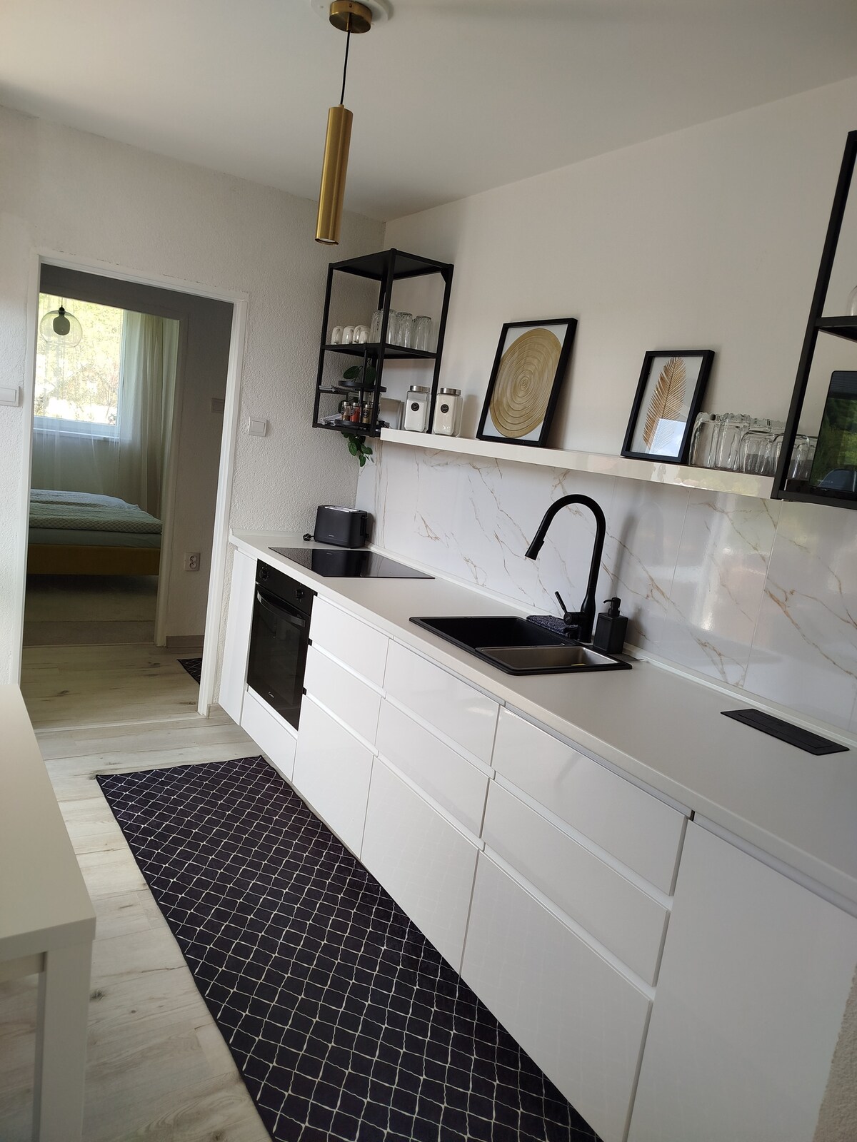 A modern kitchen is showcased with sleek white cabinetry and black accents. A stylish sink and faucet are positioned in front of a marble backsplash. Shelves display decorative items and glassware. A small dining table is visible, and a door leads to a nearby sleeping area.