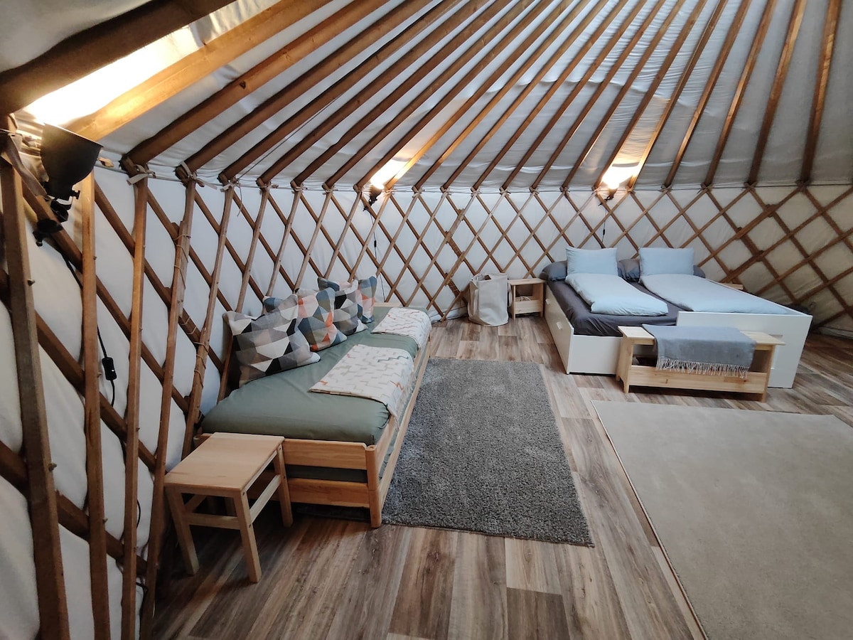 The interior of the yurt features a warm wood design with a spacious layout. A large double bed is paired with a cozy sofa bed, suitable for additional guests. Soft pillows are arranged on the sofa, with a neutral rug enhancing the inviting feel of the space.