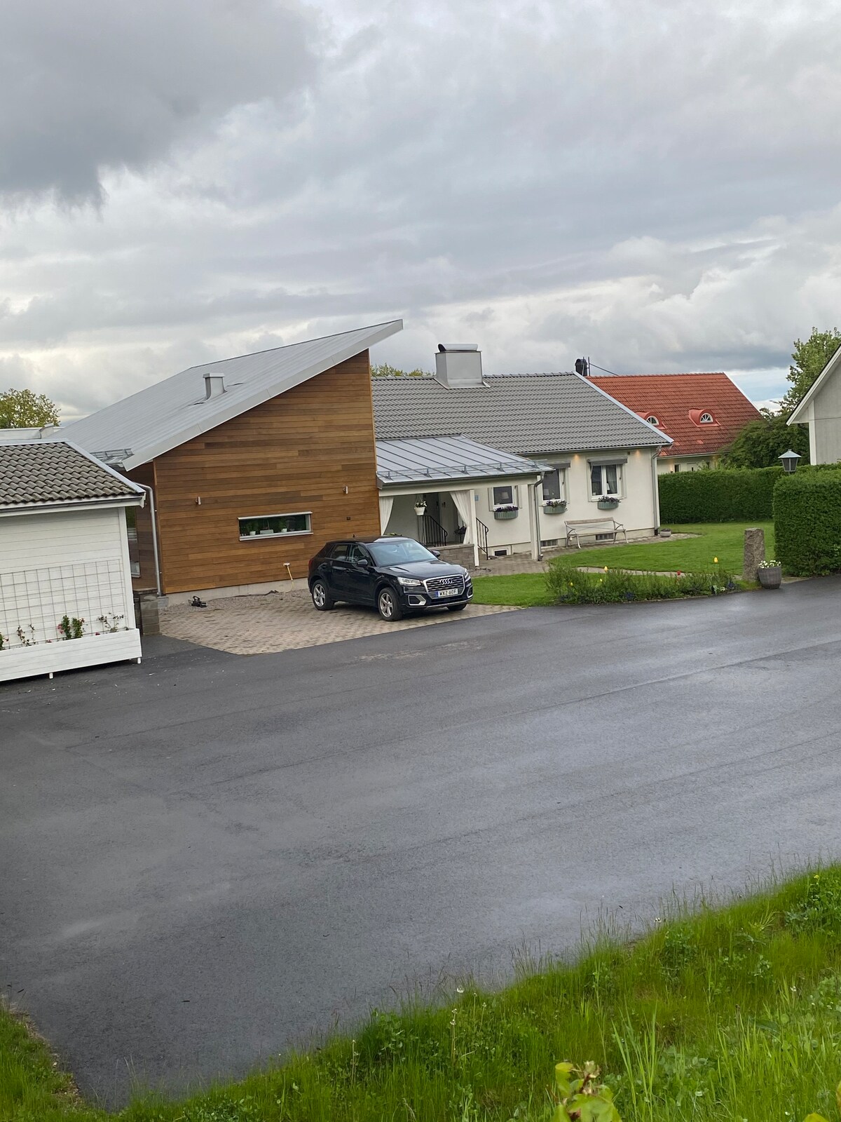 A modern house with a sloped roof features a prominent wooden façade. A paved driveway leads to the entrance, where a dark vehicle is parked. Lush green grass frames the property, and nearby houses with different roofing styles are visible in the distance.