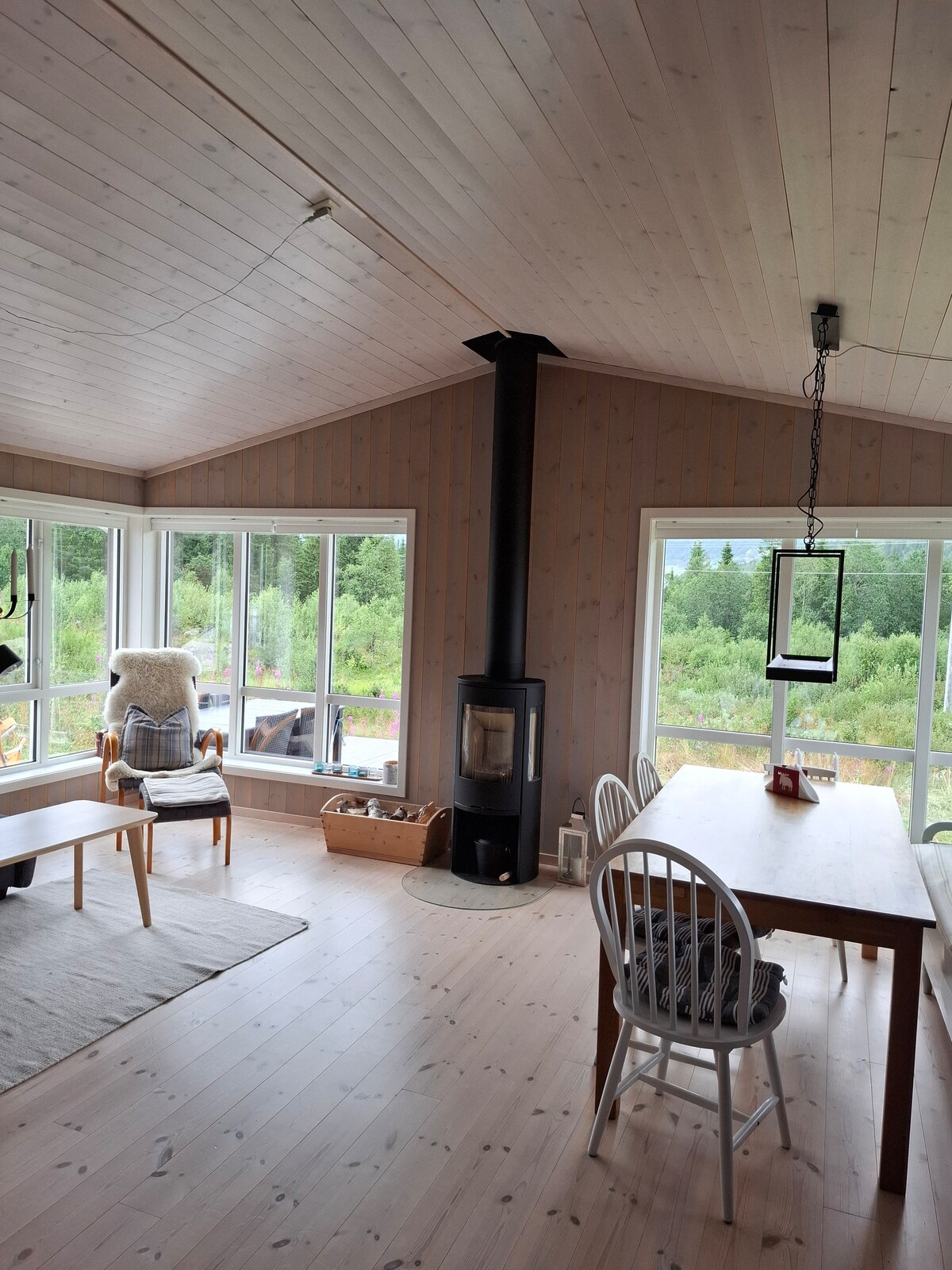 A bright living area is filled with natural light from large windows overlooking the greenery outside. A modern wood stove is positioned centrally, complemented by a dining table with chairs and a cozy bench. Soft textures create an inviting atmosphere.