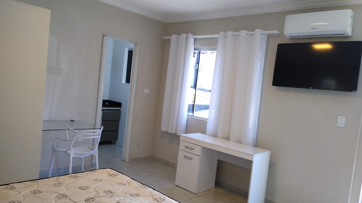 A well-lit room features a spacious bed with a patterned bedspread. A desk and chair are positioned against the wall, accompanied by a mounted television. Natural light enters through a large window dressed with sheer white curtains, providing a bright and airy feel.