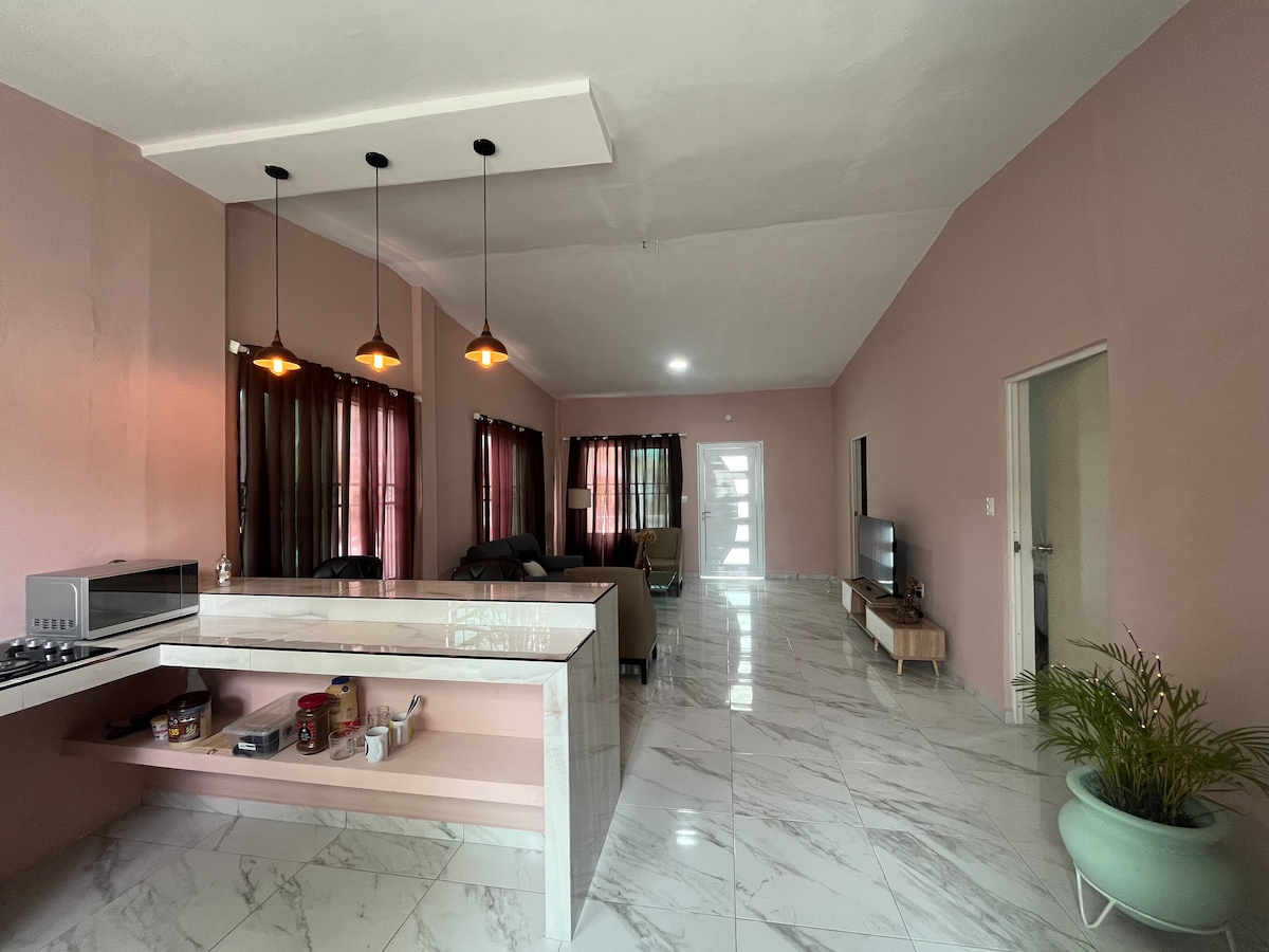 An open concept living area and kitchen are featured, with a modern island bar and contemporary lighting. The spacious lounge includes comfortable seating and a TV. Large windows allow natural light to fill the room, with lightly colored walls enhancing the overall ambiance.