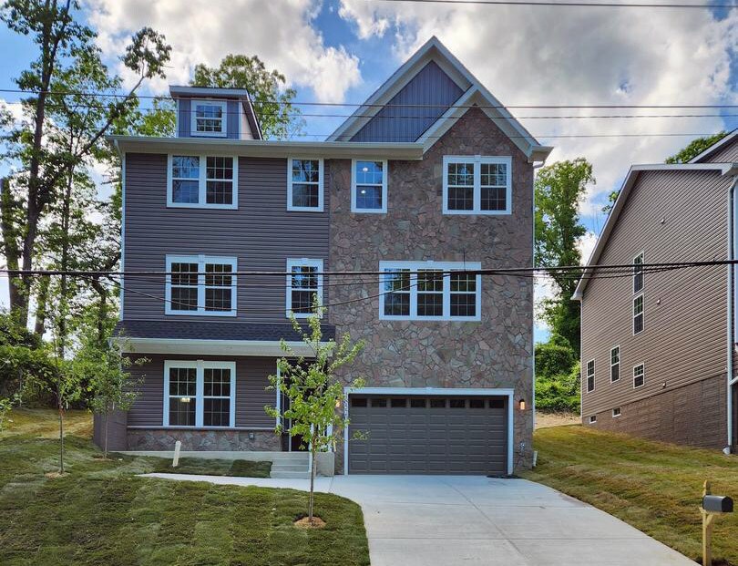 A modern three-story home features a combination of stone and siding exteriors. Large windows allow ample natural light, while a driveway leads to a two-car garage. A well-maintained lawn with small trees adds a touch of greenery to the property.