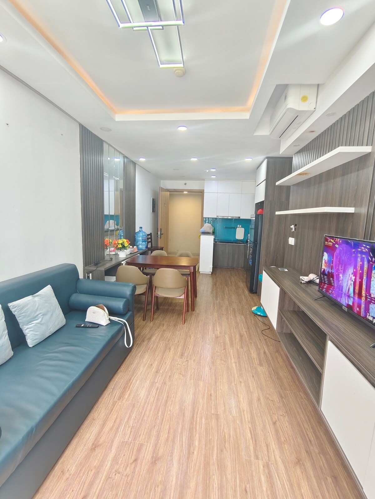 An open living area features a dark sectional sofa complemented by light-colored cushions. A wooden table with matching chairs is positioned nearby, and a television is mounted on the wall. Bright lighting illuminates the space, highlighting the modern kitchen area in the background.