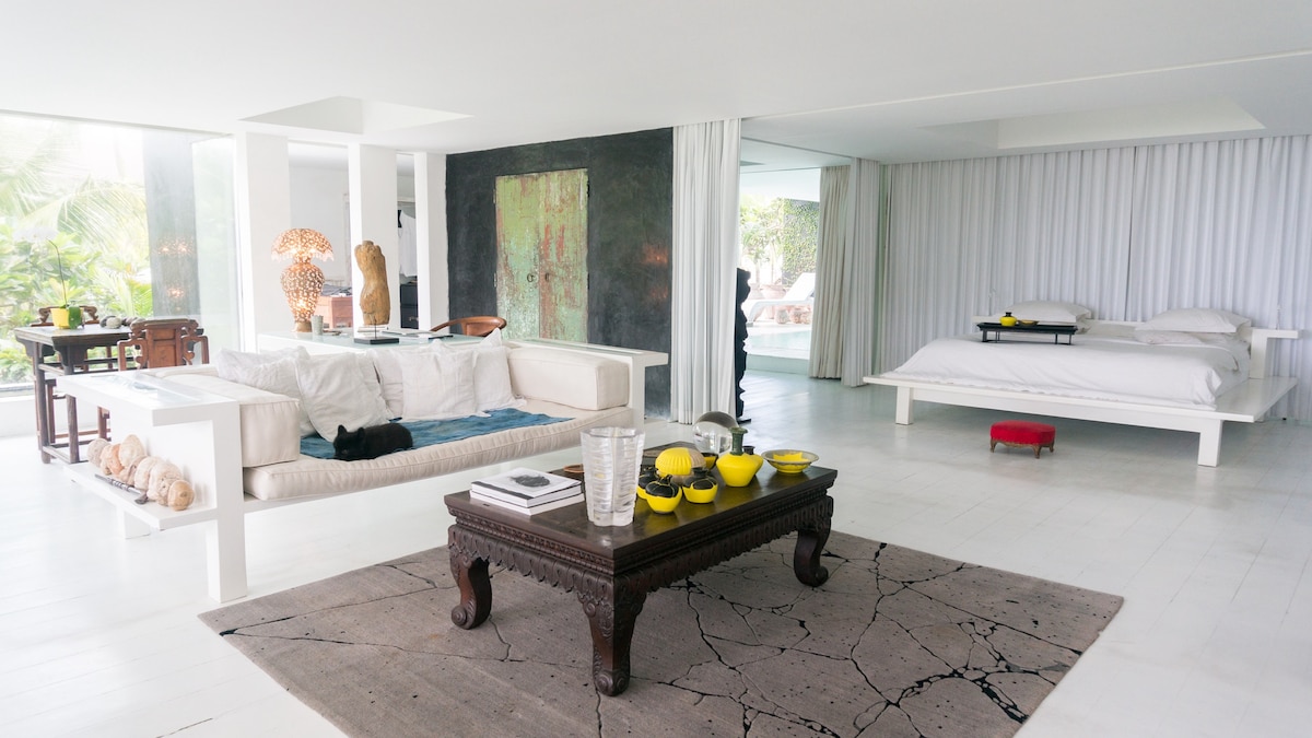 A spacious living area is highlighted by a sleek white sofa and a dark wood coffee table, adorned with various decorative items. The room features a large bed in the background, with soft linens, and natural light flooding in through expansive windows, creating an airy environment.