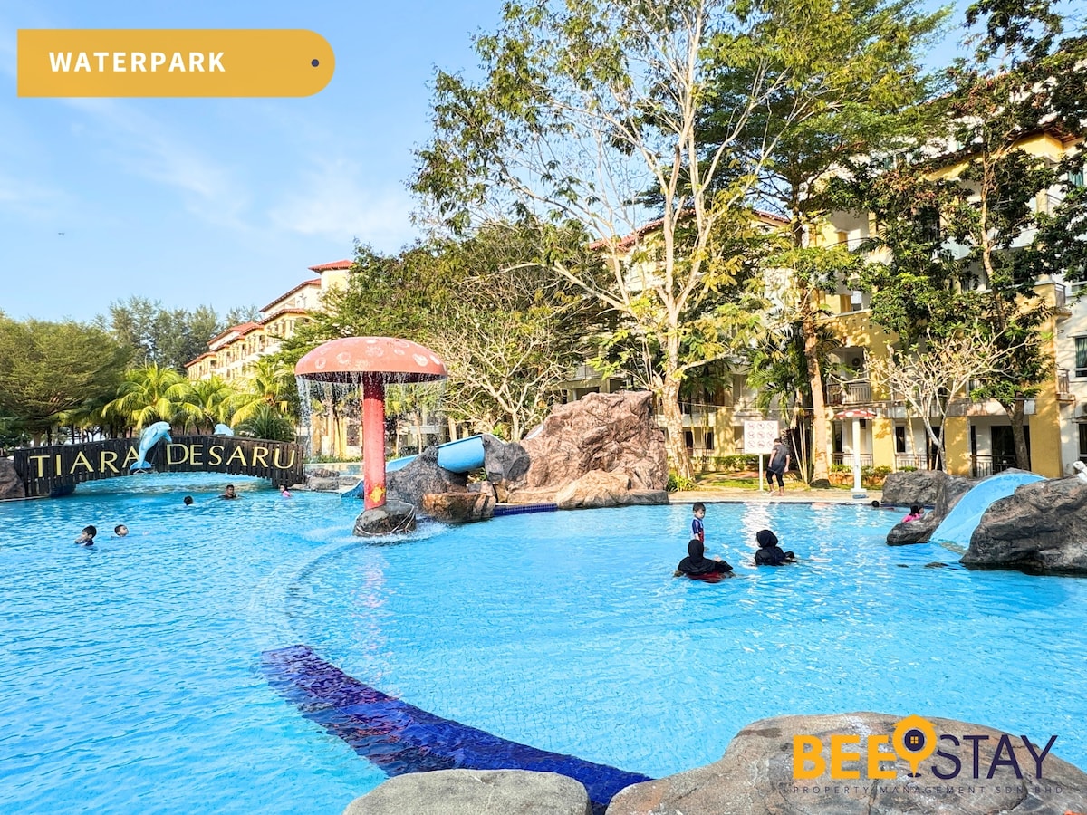 A waterpark features a large pool surrounded by lush greenery and colorful rock formations. Water slides and playful structures are visible, with guests enjoying the water. Neighboring buildings include an entrance sign labeled 'Tiara Desaru', adding to the vibrant atmosphere.