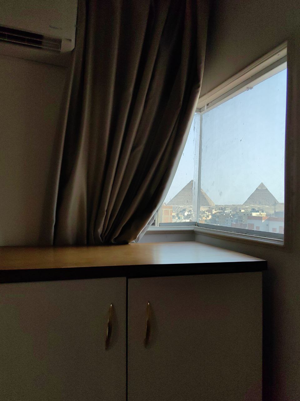 A corner of a room is depicted, showcasing a window with light filtering through. The window offers a view of pyramids in the distance, framed by sheer curtains. A minimalist cabinet with golden handles is positioned below the window, adding functional storage to the space.