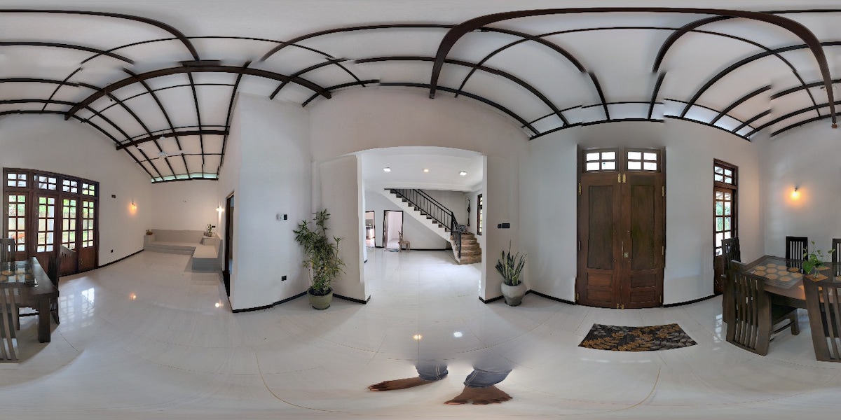 An open living space is highlighted by high ceilings and a sleek staircase leading to an upper level. Natural light pours in through multiple wooden-framed windows, illuminating the polished floors. Potted plants add a touch of greenery to the inviting atmosphere.