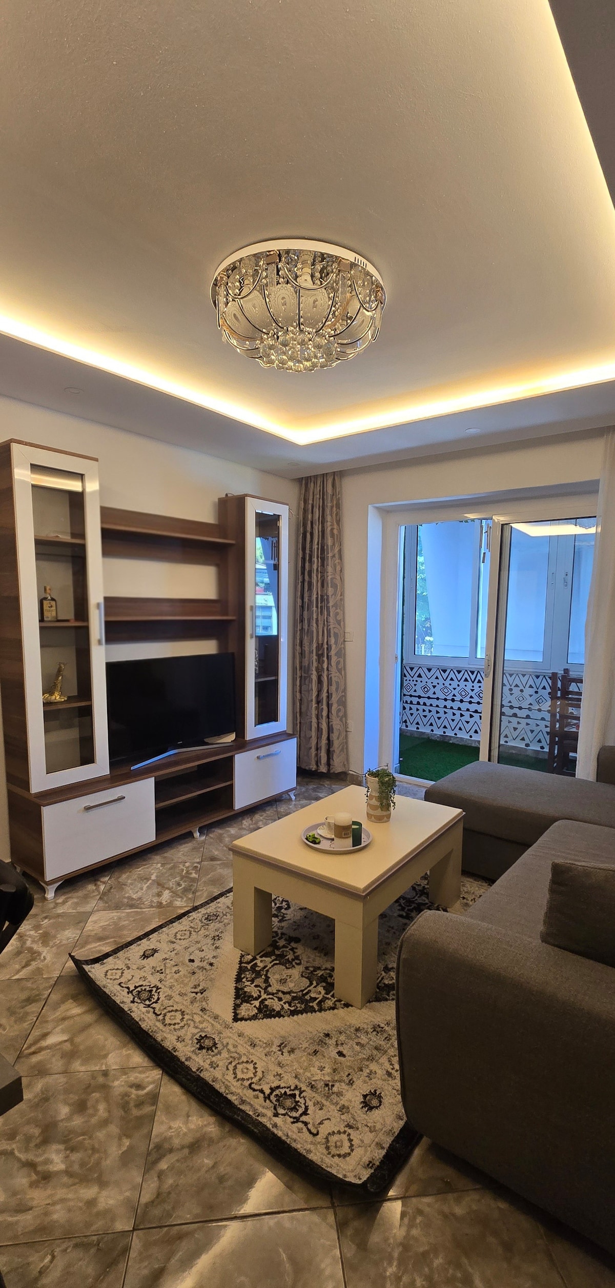 A cozy living area is featured, showcasing a modern entertainment unit with a television and decorative items. Natural light enters through glass doors, leading to an outdoor space. A light-colored coffee table sits atop a patterned rug, complemented by a gray sectional sofa.