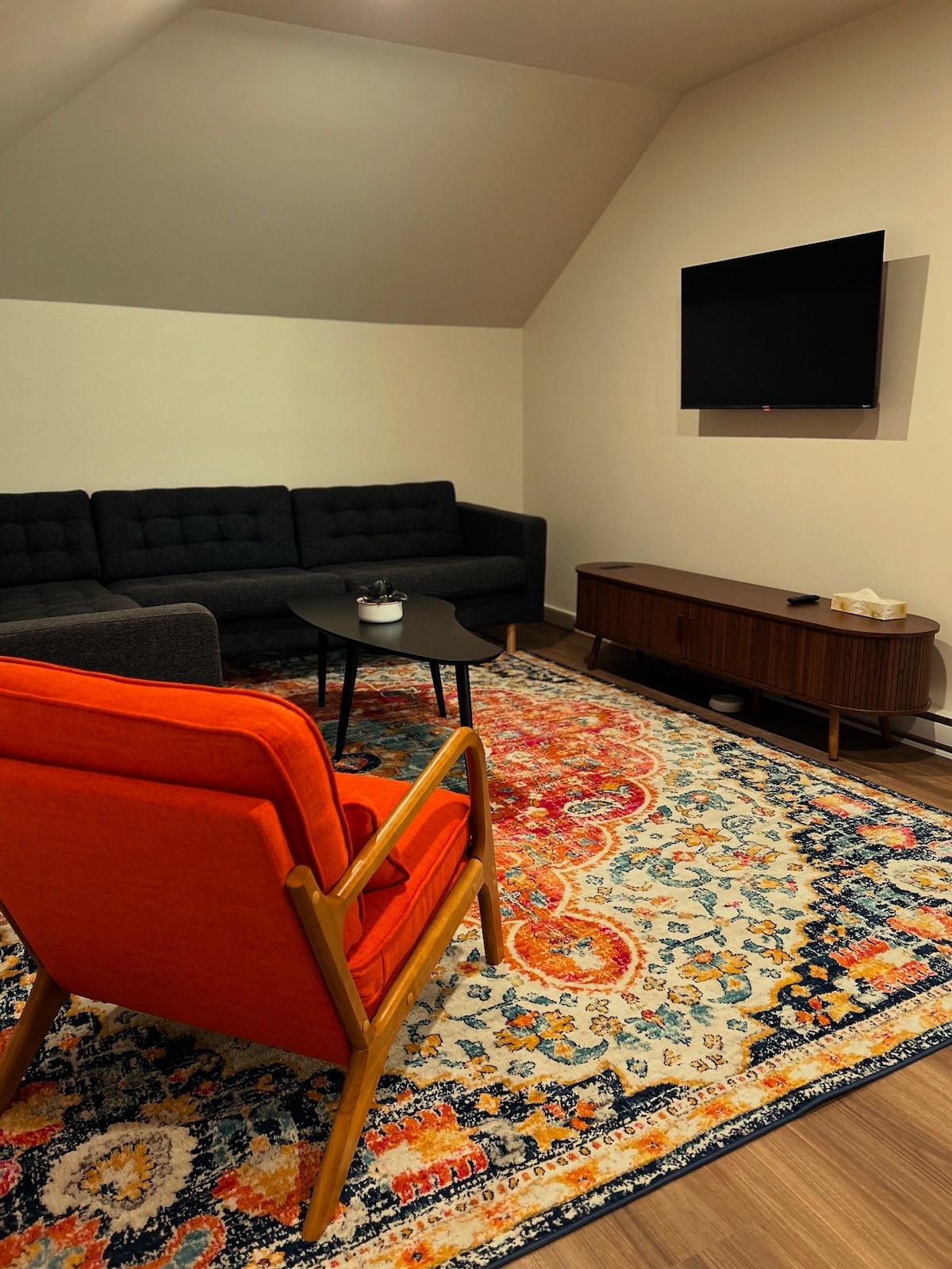 A spacious living area is highlighted by a dark sectional sofa and an orange accent chair, positioned on a patterned rug. A flat-screen TV is mounted on the wall above a stylish media console, creating a cozy and functional entertainment space.