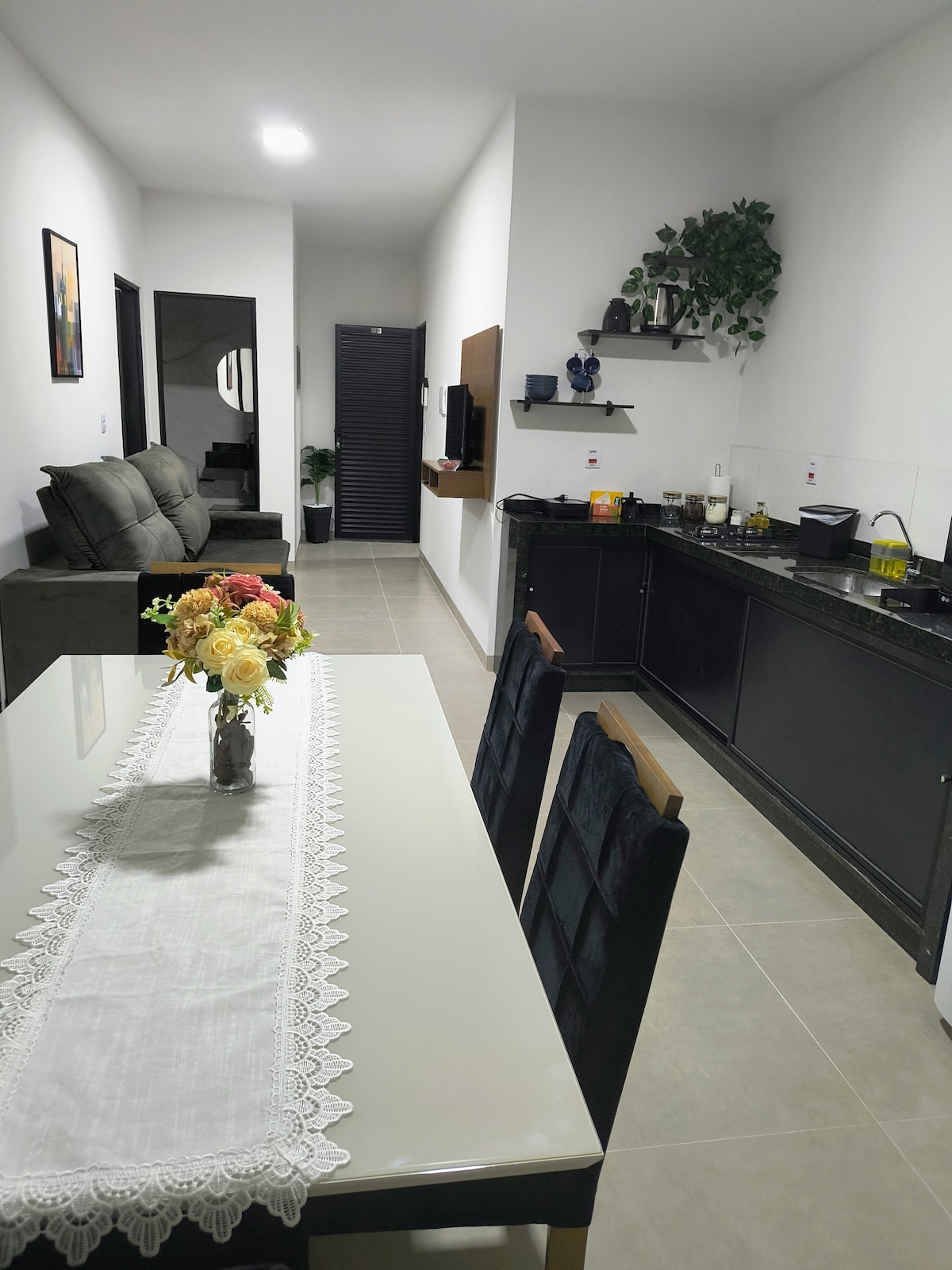 A neat dining area is presented with a table set for four, adorned with a floral centerpiece. The adjacent kitchen features dark cabinetry and offers modern appliances. A comfortable seating arrangement is visible in the background, enhancing the space's functional atmosphere.