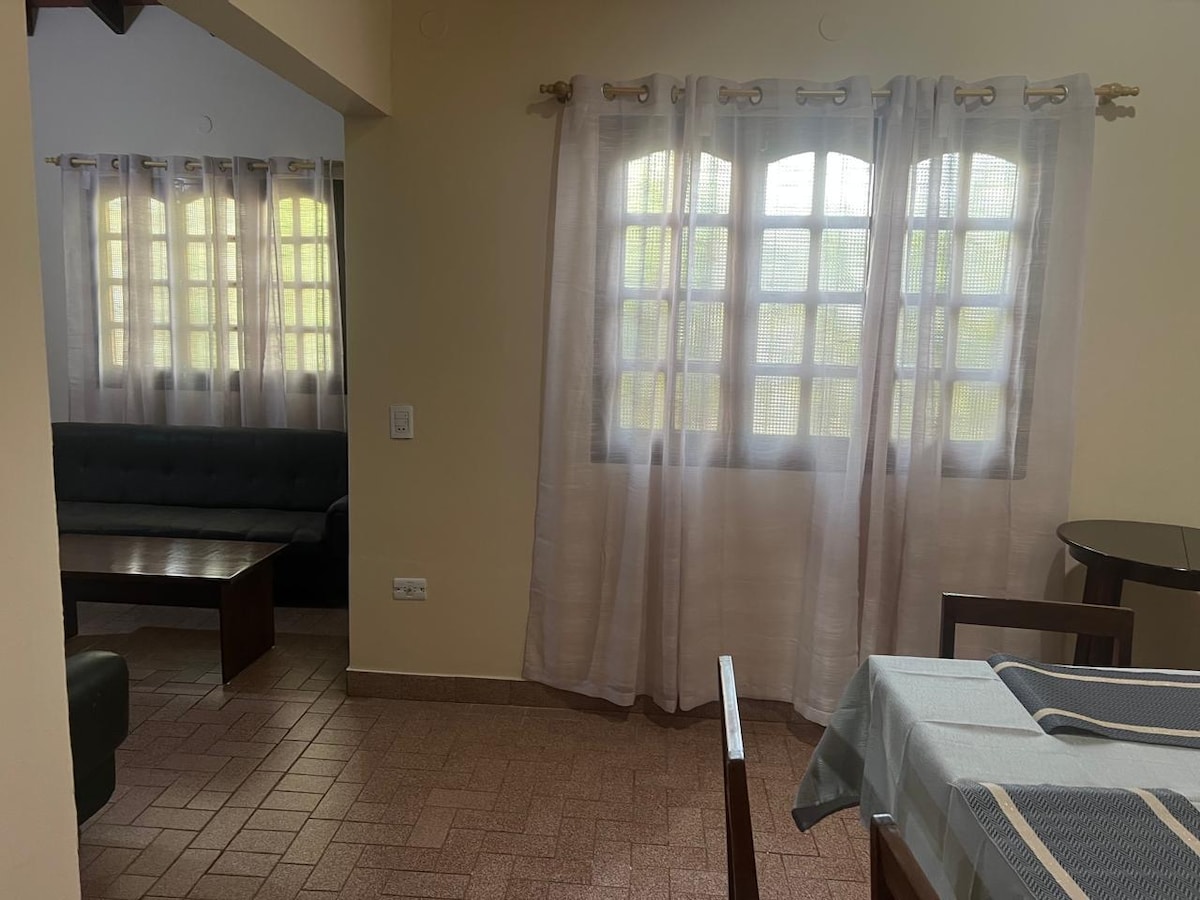 A cozy living space is presented with a dark sofa against one wall and a round dining table positioned nearby. Soft, sheer curtains frame the large windows, allowing natural light to fill the room. The flooring is tiled in a warm tone, enhancing the inviting ambiance.