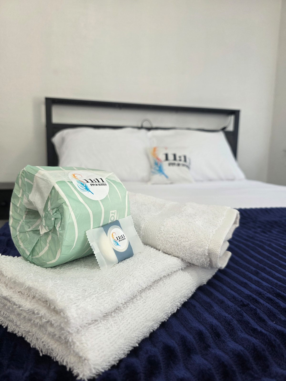 A neatly arranged bed is presented with a white quilt cover and decorative pillows. Fresh towels are stacked beside a rolled towel, both in white, along with a personal item placed nearby, emphasizing comfort and convenience.