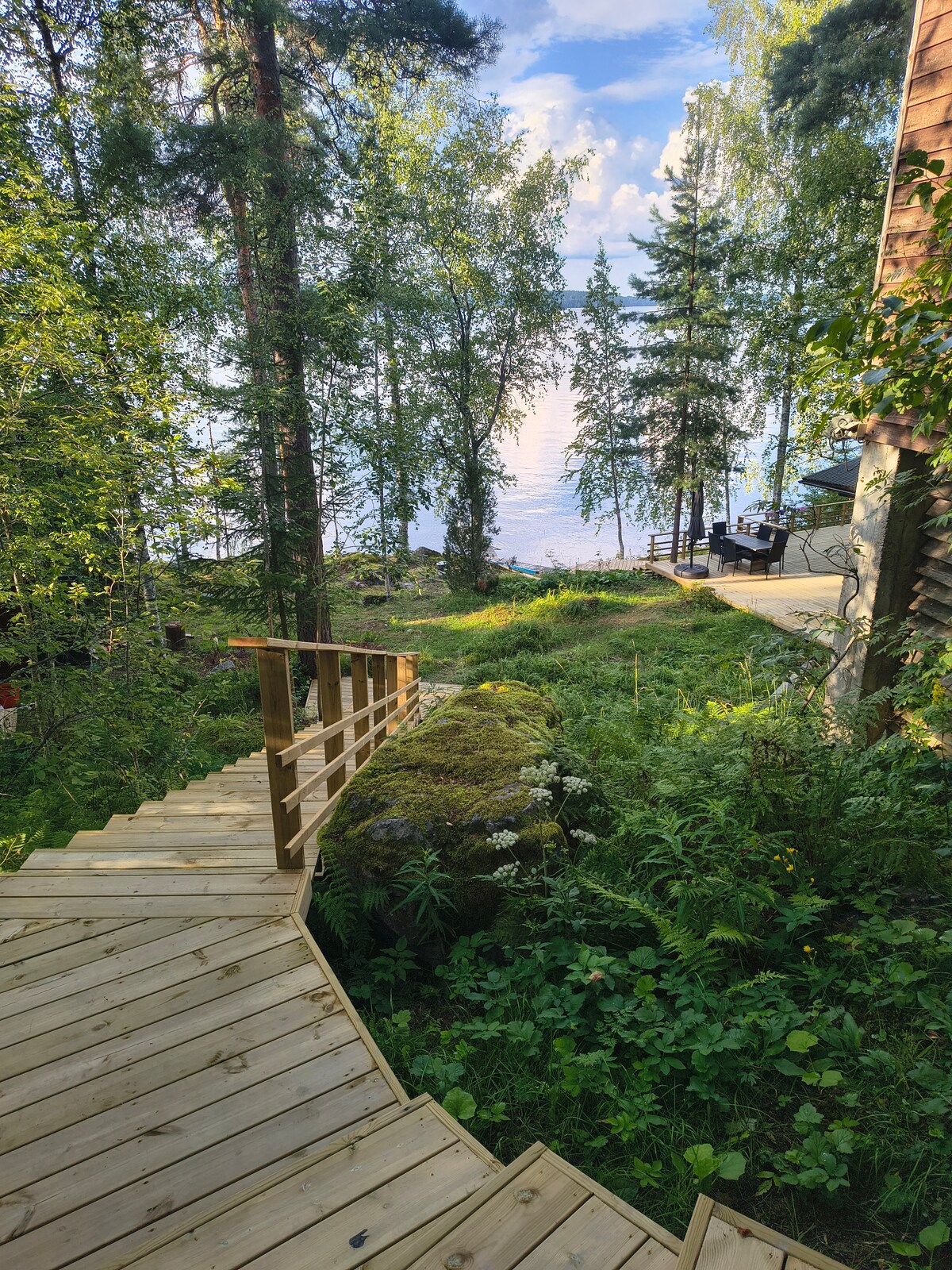 A winding wooden staircase descends through lush greenery towards a serene lake. Tall trees and vibrant foliage frame the path, while the water glimmers softly in the background. A small dock is visible along the lakeshore, inviting relaxation in nature.