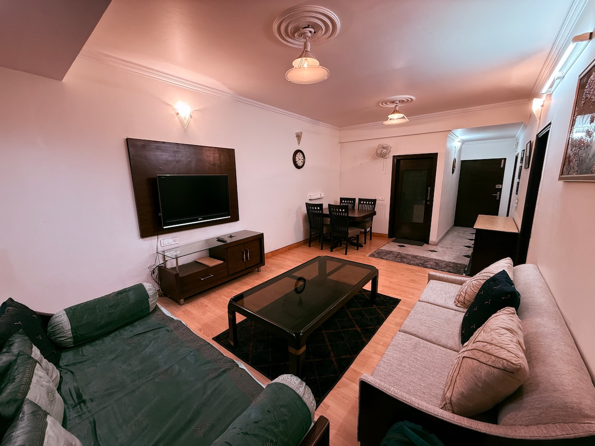 A spacious living area is depicted, featuring a comfortable sofa with an inviting green throw. A coffee table sits centrally, complemented by a flat-screen TV mounted on the wall. A dining area is visible in the background, providing a cozy space for gatherings.