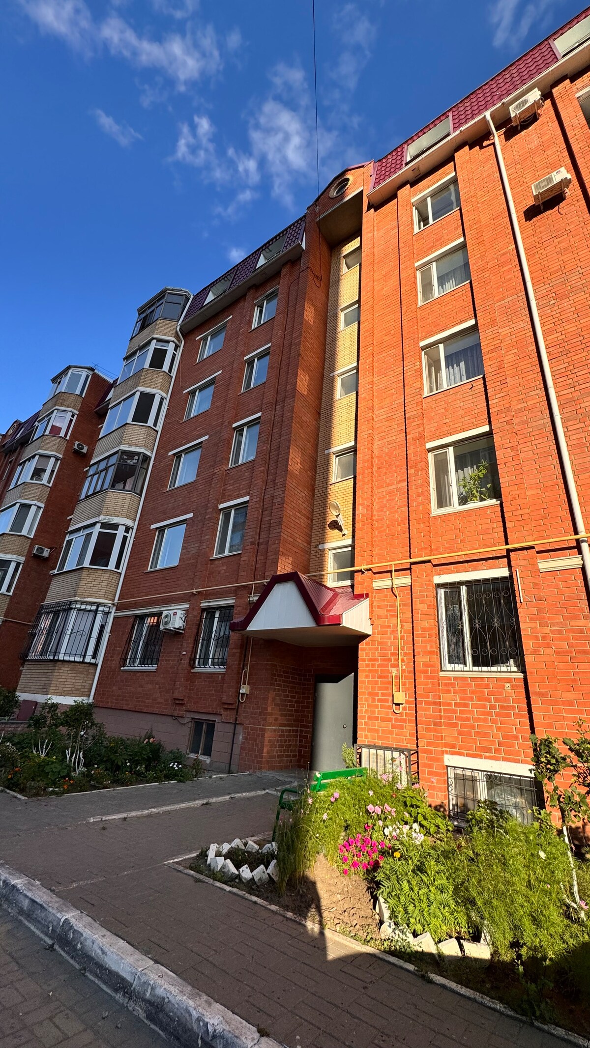 The exterior of a five-story brick building is presented, featuring large windows and a sloped roof. A small entranceway is highlighted by a red awning. The well-kept garden displays various plants and flowers along the pathway leading to the entrance.
