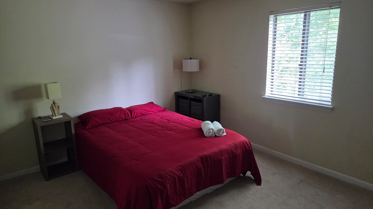 A well-lit bedroom features a spacious bed with a red coverlet. A pair of white towels is neatly arranged on top of the bed. A small nightstand holds a minimalist lamp, while a window with blinds allows natural light to illuminate the room.