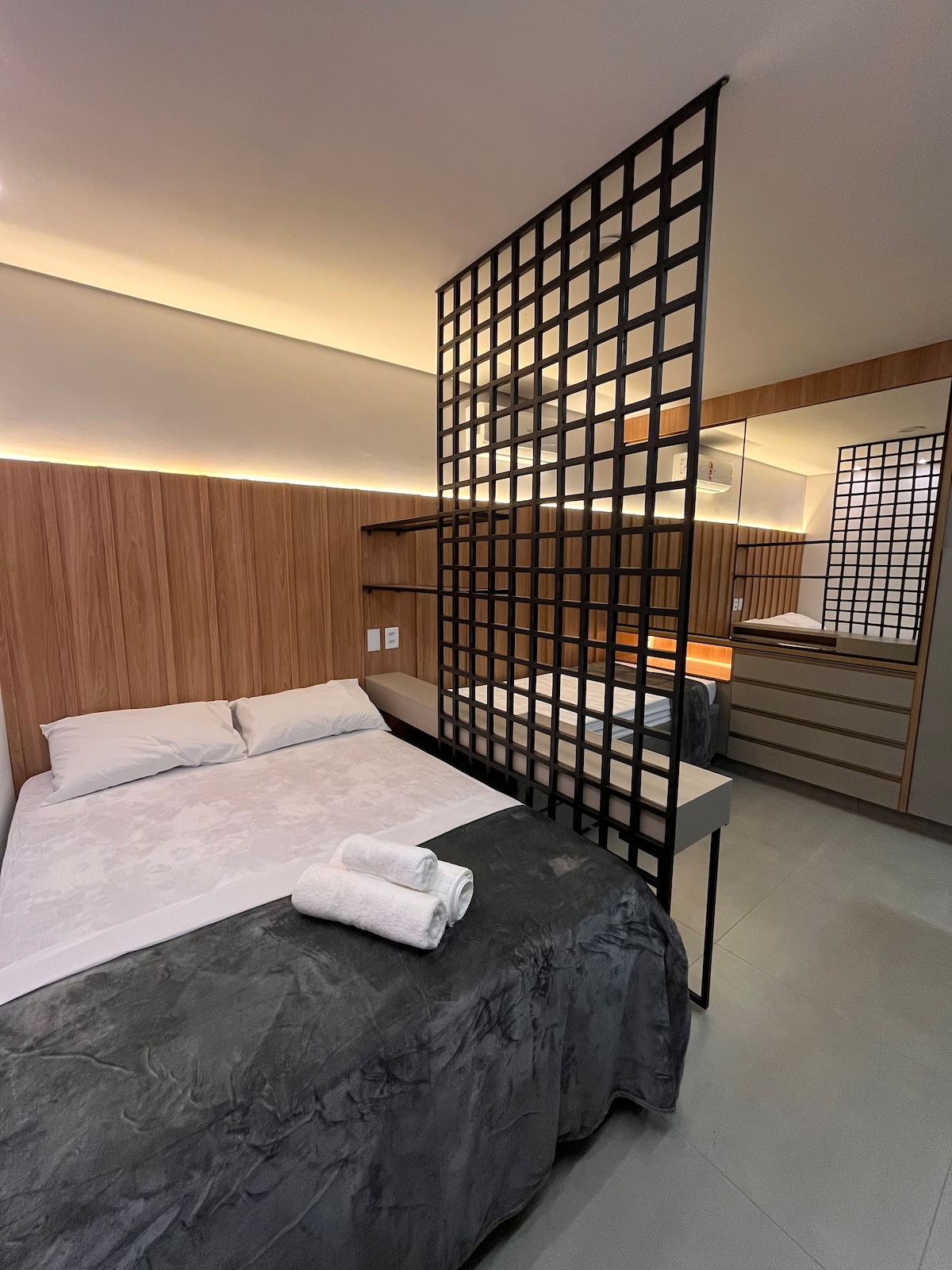 A modern bedroom features a spacious bed with crisp linens and soft towels folded at the foot. A stylish divider adds privacy, while warm lighting enhances the wood-paneled walls. A mirrored wardrobe and minimalist furnishings are noticeable, providing a clean, functional space.