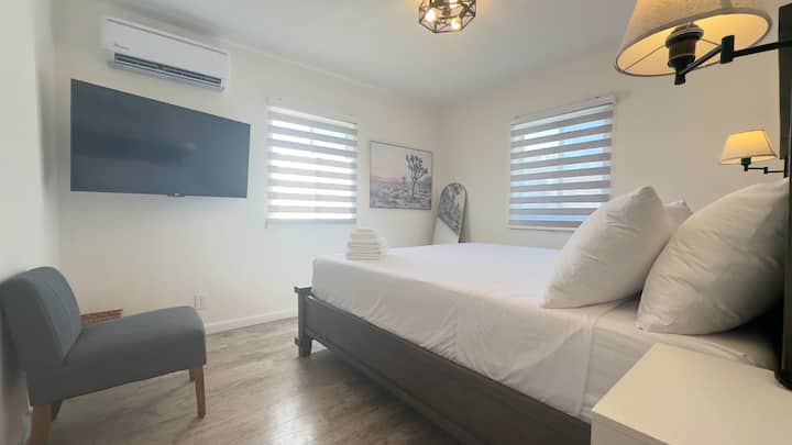 غرفة نوم رئيسية تتميز بسرير كاليفورنيا كينج وتلفزيون ذكي ومصابيح جانبية أنيقة وكرسي منجد رمادي مريح — مصمم للراحة والأسلوب الصحراوي العصري.