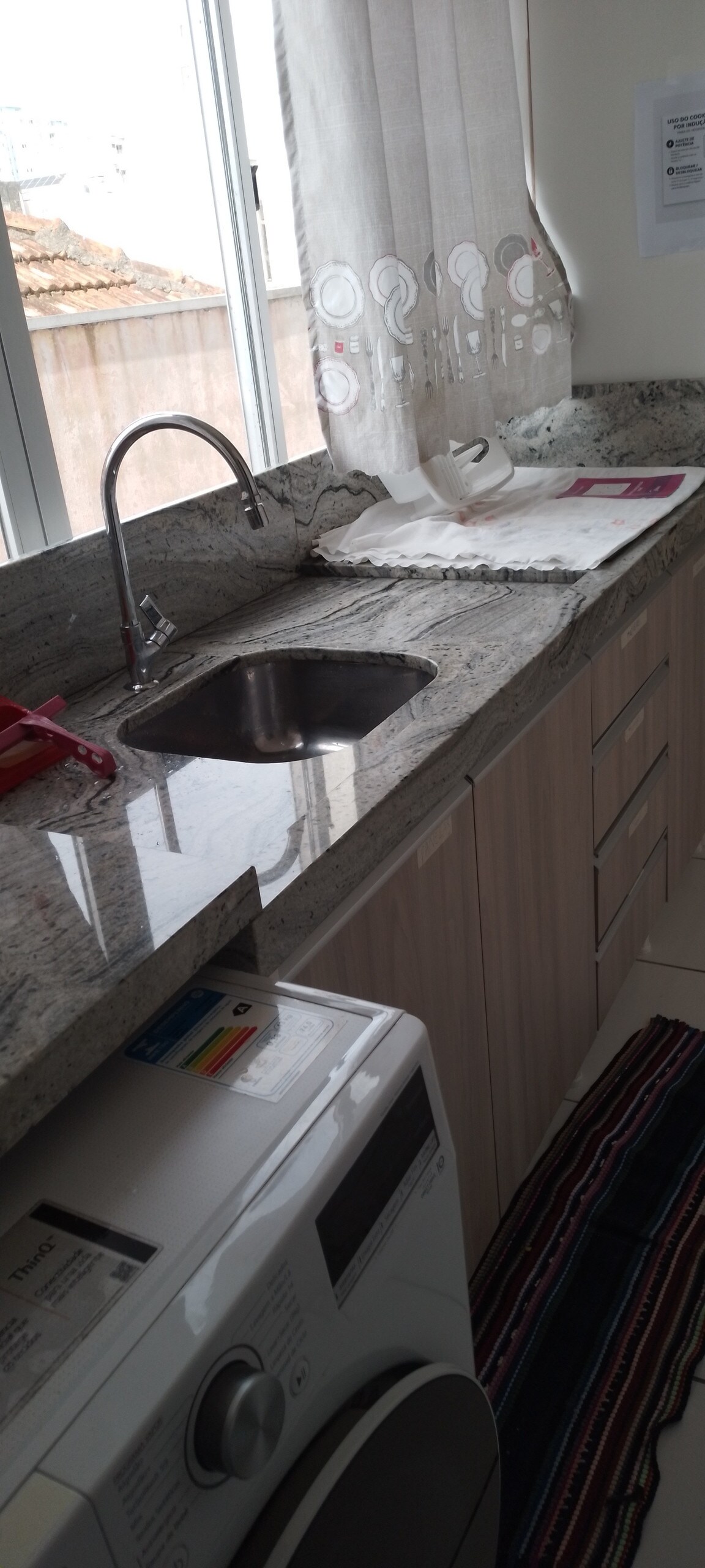 The kitchen area is equipped with a sleek granite countertop and a small sink, complemented by wooden cabinetry. A washing machine is visible beneath the counter, while natural light enters through a nearby window adorned with sheer curtains.