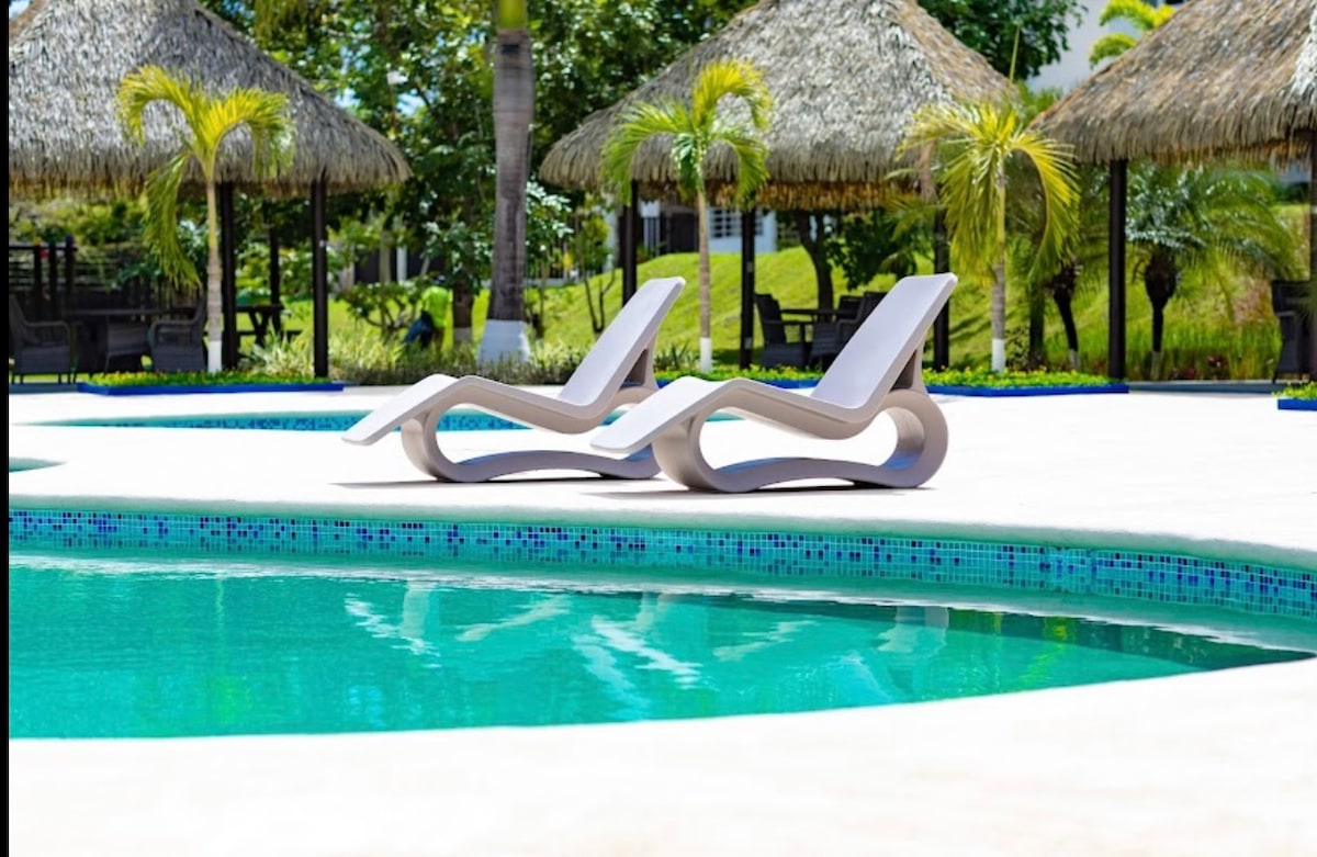 Two modern lounge chairs are positioned near a shimmering turquoise pool, surrounded by lush greenery and tropical palm trees. Thatched-roof cabanas offer shaded seating areas nearby, creating a tranquil environment for relaxation.