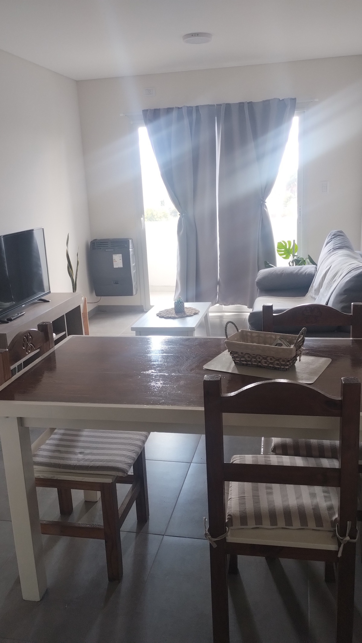 The living area features a wooden dining table surrounded by chairs and a contemporary couch near a large window. Natural light enters through the partially opened curtains, highlighting a television unit and a small decorative plant beside the window.