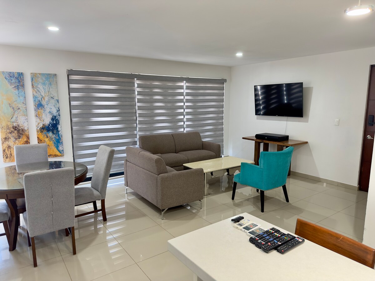 A spacious living area is visible, featuring a gray sofa and a light coffee table. Two dining tables are present, one with gray chairs and another with wooden seating. A wall-mounted TV is positioned across from the sofa, and large windows are adorned with striped shades.