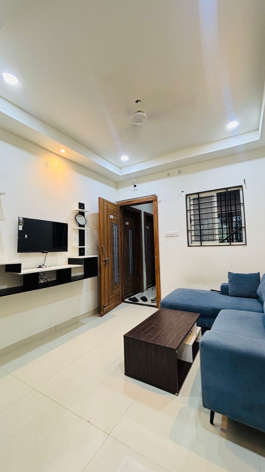 A cozy living space features a blue sectional sofa and a low wooden coffee table. A wall-mounted television is positioned above a minimalistic shelf. Natural light enters through a window adorned with a security grill, and the entrance door is partially open, revealing a glimpse of the hallway.
