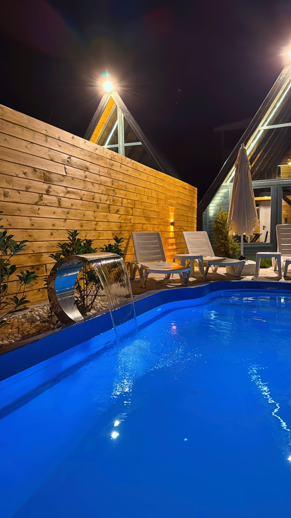 A serene private pool is illustrated, featuring a gentle waterfall feature that adds a tranquil sound. Light blue water is surrounded by white lounge chairs. A wooden fence and landscaped greenery provide a cozy enclosure, enhancing the peaceful atmosphere of the outdoor space.