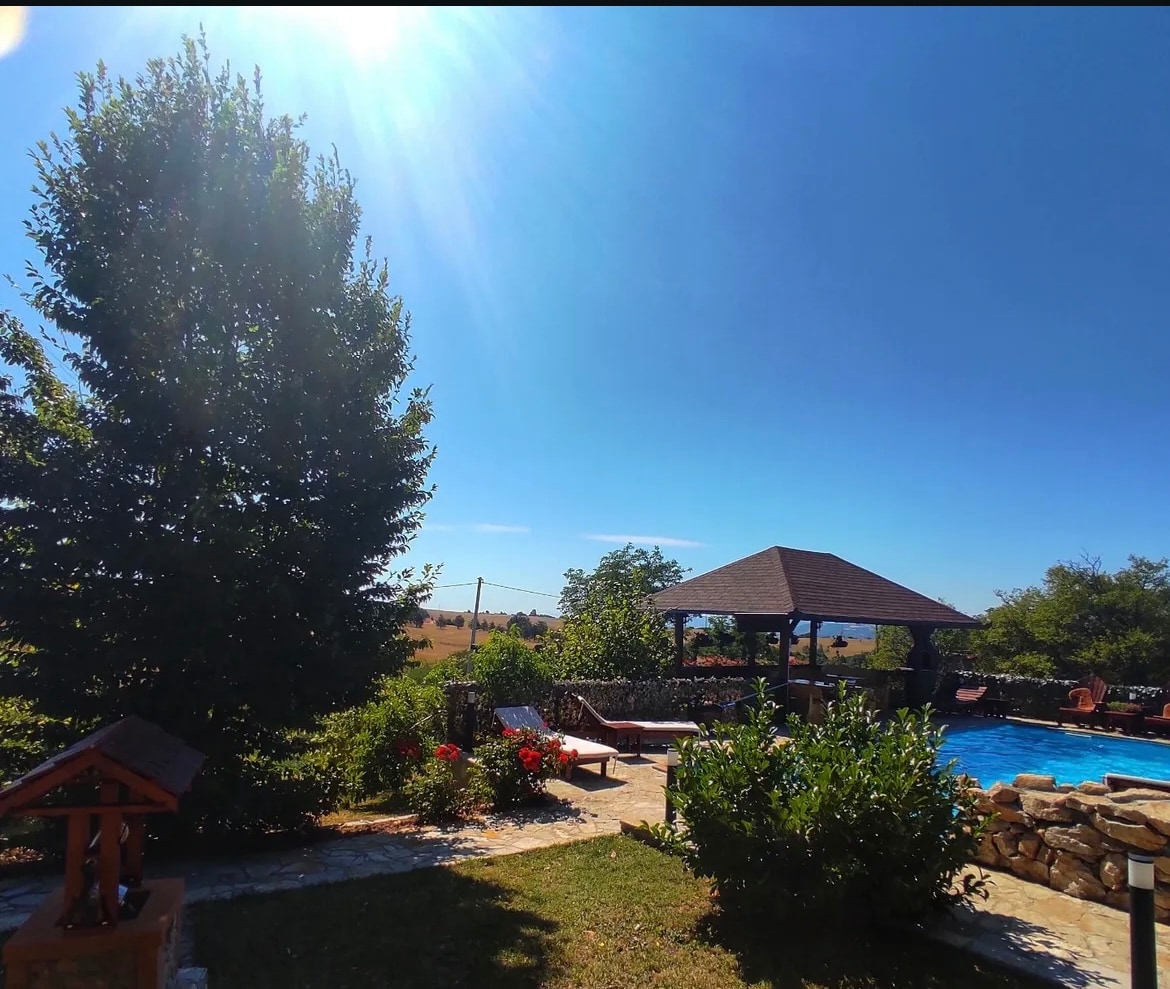 The image showcases a sunlit outdoor area with lush greenery, featuring a pool surrounded by lounge chairs and a shaded gazebo. Bright flowers add color to the landscape, enhancing the natural setting. The clear blue sky suggests a warm day.