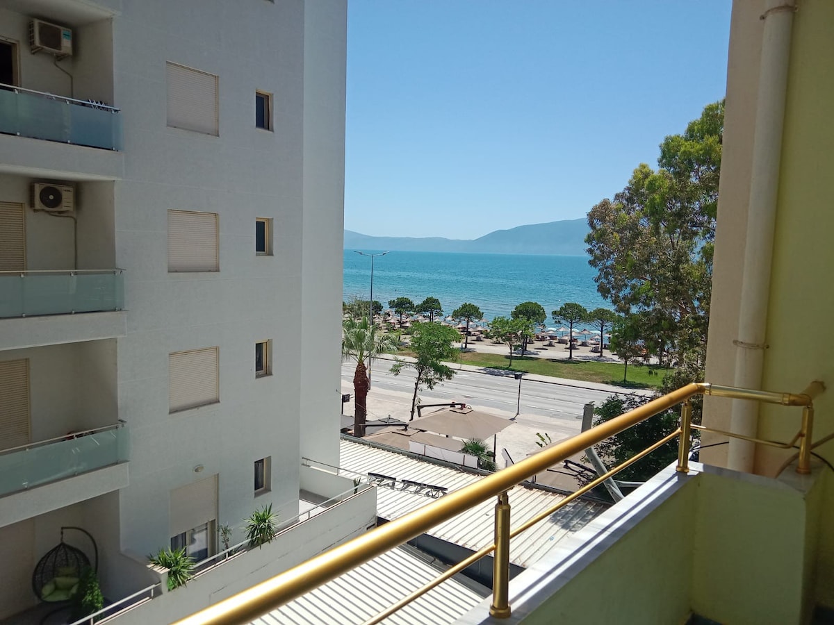 A balcony view overlooks the beach and ocean, with gentle waves visible in the distance. Palm trees line the shore, and a coastal road is positioned below. The scene is framed by a white building on the left and bright blue sky above.