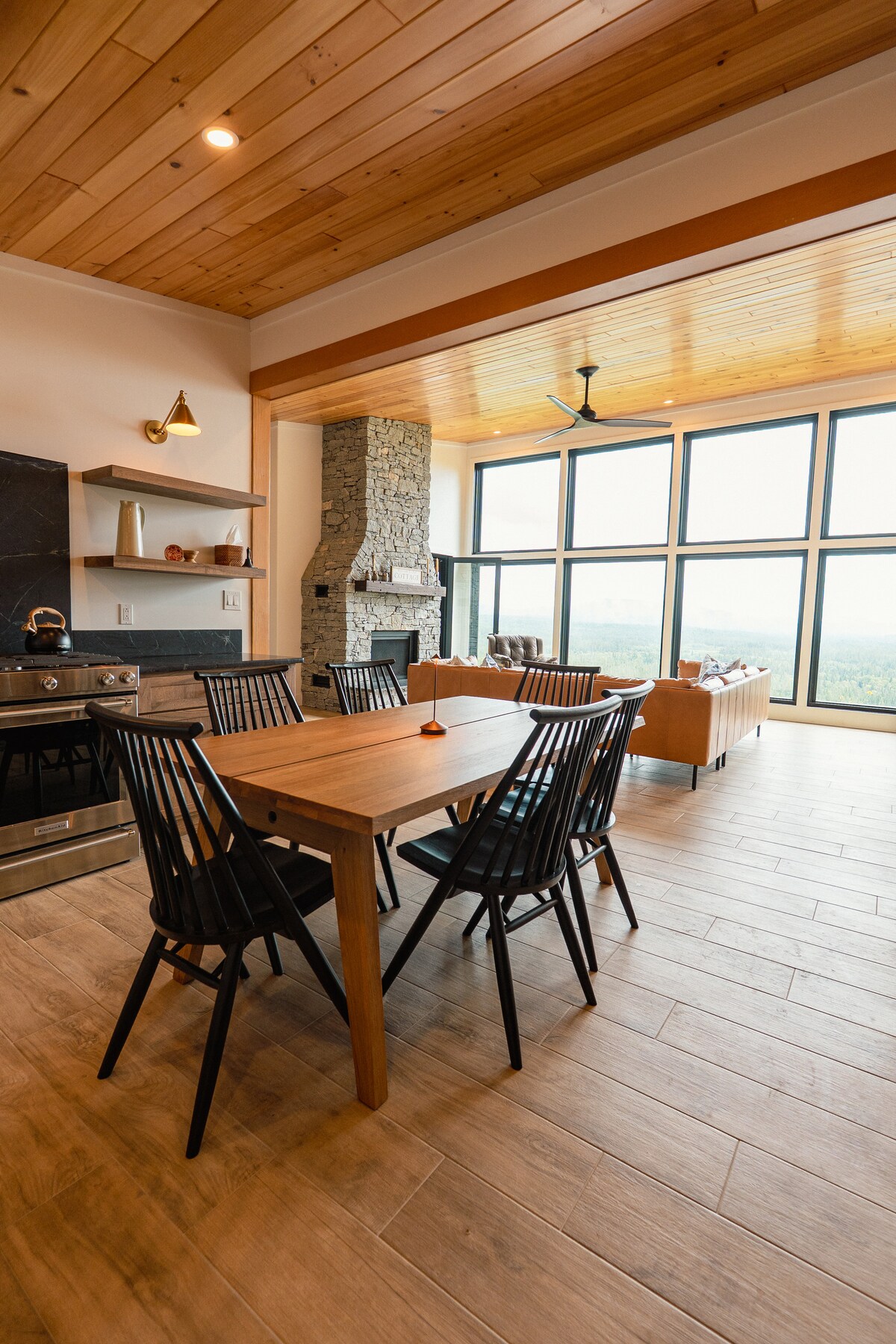 A modern dining area is prominent, featuring a wooden dining table surrounded by six black chairs. Floor-to-ceiling windows provide expansive valley views, while a stone fireplace adds a touch of warmth. Simple decor and natural light enhance the inviting ambiance of the space.