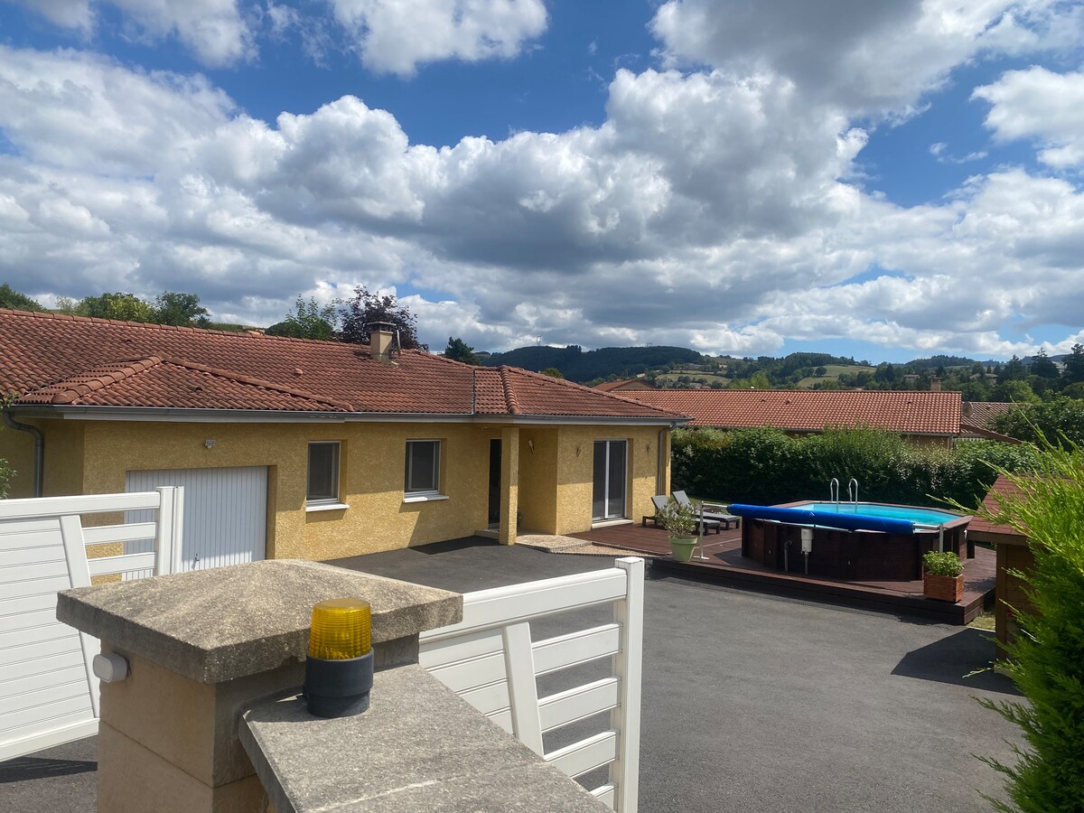 A single-story house displays a warm yellow exterior, with a spacious driveway leading to a private area. A swimming pool is visible in the background, surrounded by greenery. Soft clouds drift across a blue sky, adding to the serene atmosphere.