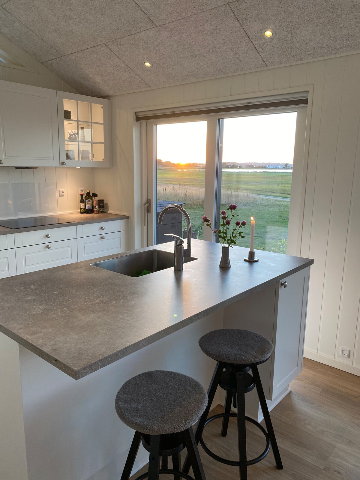 A modern kitchen features a sleek countertop with a spacious sink. Two bar stools are positioned around the island. A window showcases a sunset over the fields, adding a serene backdrop. A small vase with flowers and a candle enhance the space.