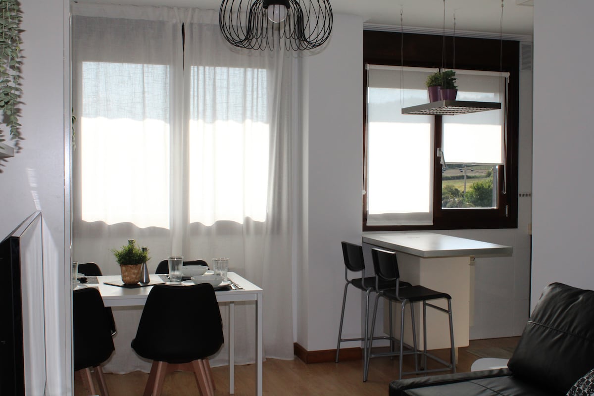 A bright living area features a dining table with four black chairs. An island counter with stools is positioned nearby. Natural light filters through sheer curtains covering large windows, contributing to an airy ambiance. A modern light fixture hangs above the dining area.