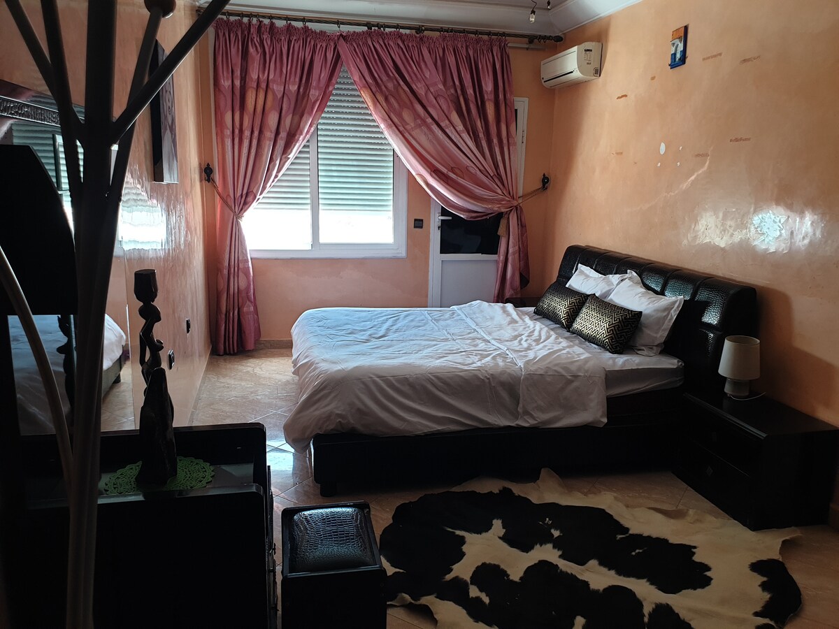 A spacious bedroom features a large bed with a simple white duvet and decorative pillows. Soft pink curtains frame the windows, allowing natural light to filter in. A patterned area rug lies on the floor, complementing a stylish black nightstand and decor elements.