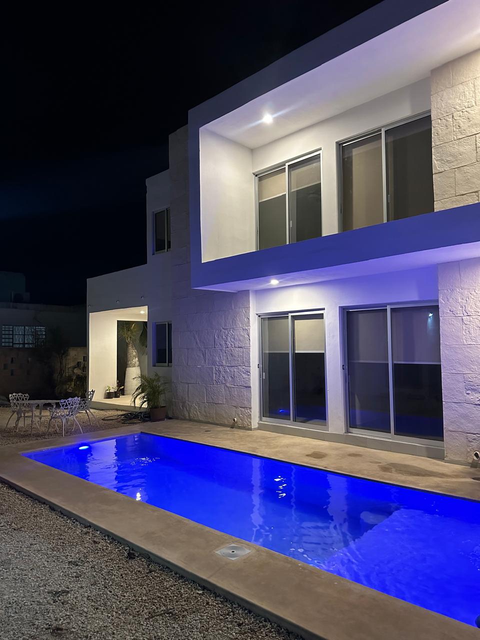 A modern two-story building is illuminated at night, showcasing expansive glass doors that reflect the soft blue light of the pool. A small patio area with outdoor seating is visible beside the shimmering water, enhancing the outdoor aesthetic.