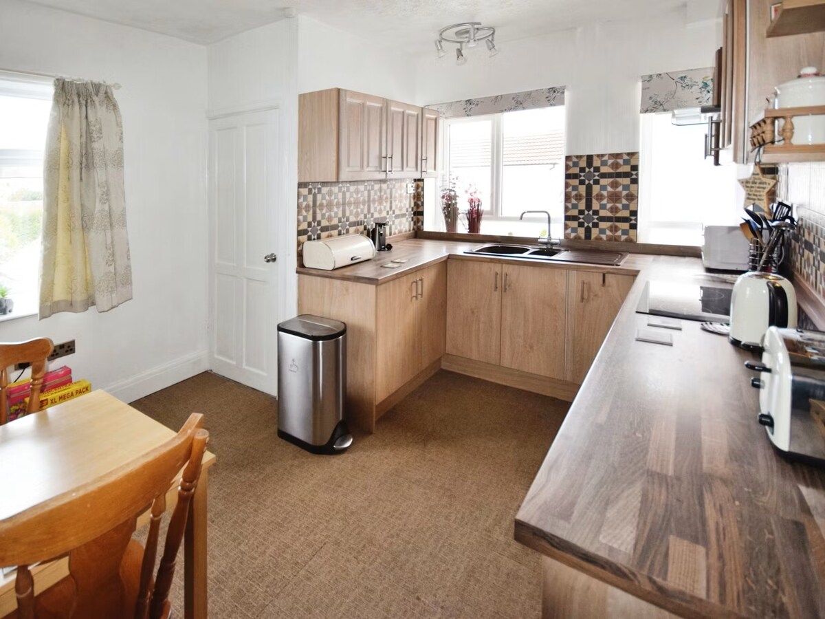 The kitchen features light wood cabinetry and a spacious countertop. A sink is positioned near the window, allowing natural light in. A small dining table with two chairs is visible, along with essential appliances including a refrigerator and a trash bin.