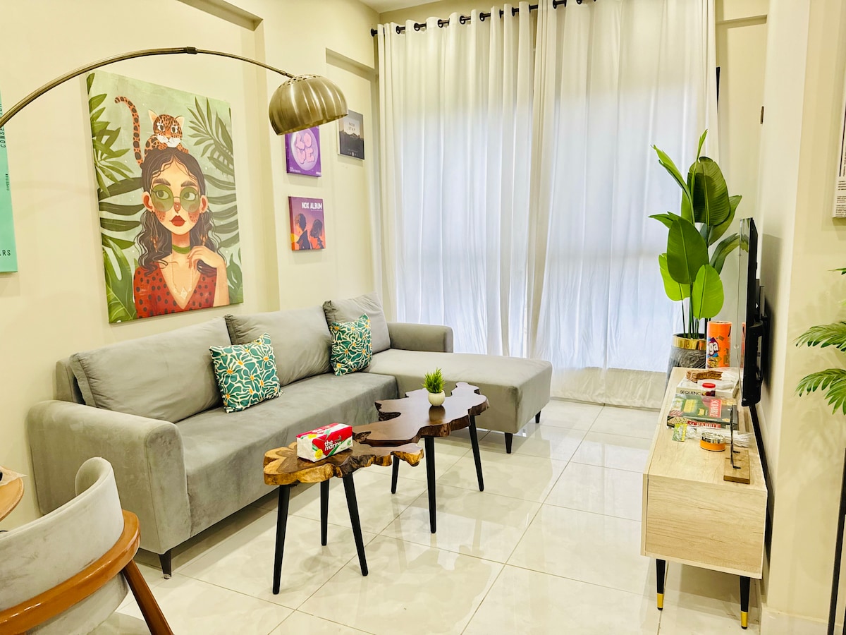 A cozy lounge area features a stylish gray sofa adorned with colorful cushions. A natural wood accent table sits in front of the sofa, complemented by decorative plants. Light-filtering curtains frame a large window, creating a bright and inviting atmosphere for relaxation.