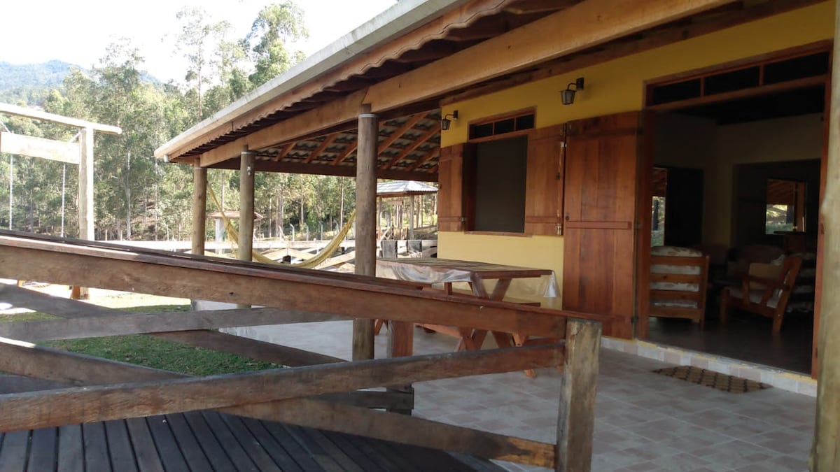 A spacious outdoor area is framed by wooden structures, featuring a charming covered porch with wooden beams. A set of comfortable seating is visible through open windows. Lush greenery surrounds the property, offering a peaceful connection to nature.