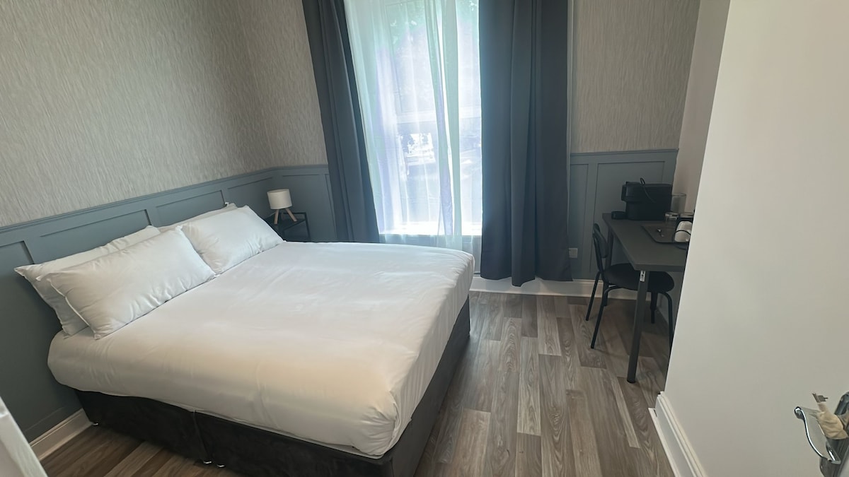 A well-lit bedroom is furnished with a spacious bed dressed in crisp white linens. A small desk and chair are positioned nearby, while soft curtains frame a window that allows natural light to fill the space. Neutral walls complement the wood flooring.