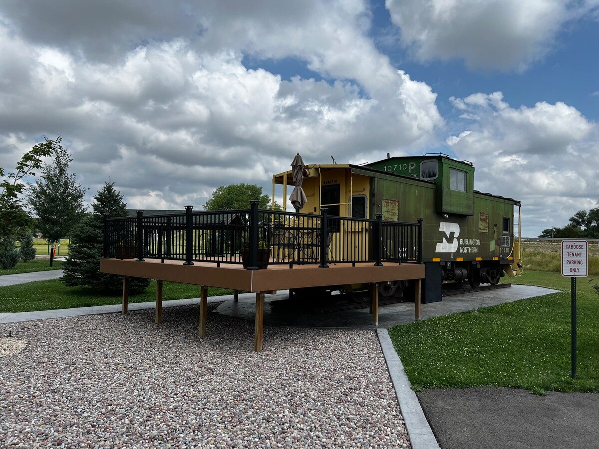 An exterior view of a remodeled train caboose, set on a wooden deck with black railings. Surrounding greenery and trees are visible, with a gravel path leading to the caboose. The sky overhead features a mix of clouds, providing a serene ambiance.
