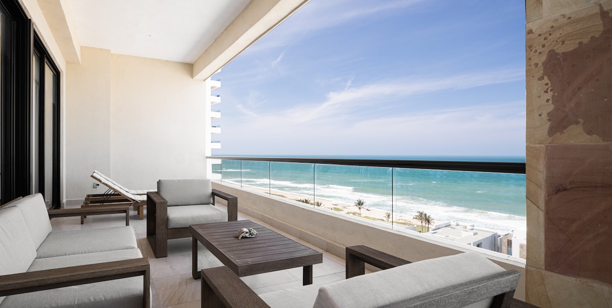 A spacious outdoor seating area is showcased, featuring comfortable sofas and a wooden coffee table. A clear view of the ocean is provided, with gentle waves visible against the sandy shore. The sky is bright and partly cloudy, adding to the serene setting.