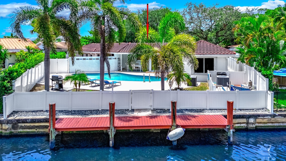 A spacious backyard is enclosed by a white fence, featuring a heated pool surrounded by palm trees. A red wooden dock extends into the water, providing access to serene canal views. Two gas BBQ grills are positioned on the patio, perfect for outdoor cooking.