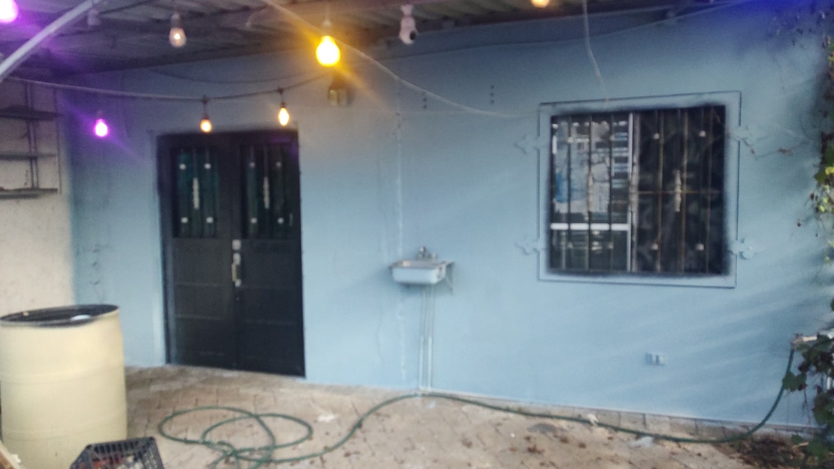 An exterior wall painted in soft blue features two black doors flanked by a window with security bars. A small wall-mounted sink is visible, along with a hose coiled on the ground. Colorful lights hang above, adding a vibrant touch to the space.