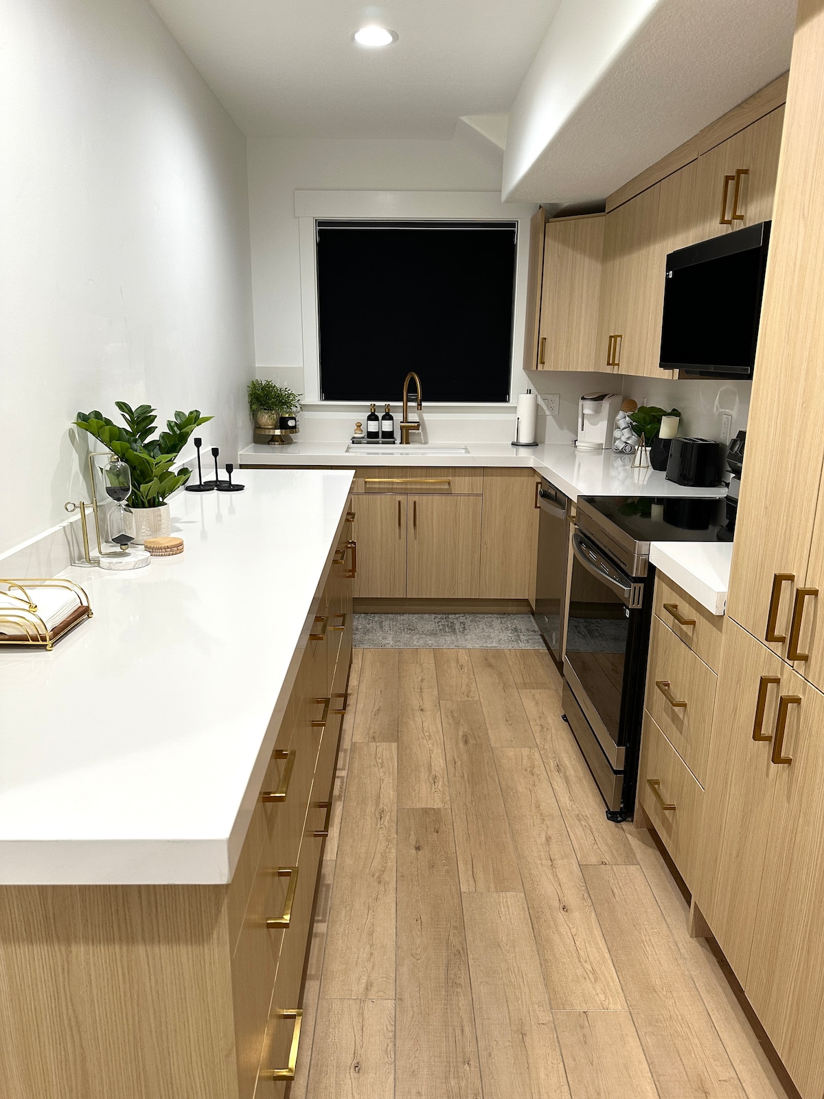 A modern kitchen is displayed, featuring light wood cabinetry with gold hardware. A white countertop extends along one side, complementing the matching backsplash. A black window treatment controls natural light, and small plants are positioned on the counter for a touch of greenery.